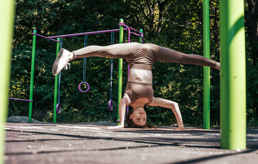 Young woman performs a headstand and twine at the same time on a sports field in the park.