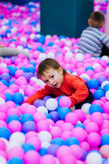 Obraz na płótnie Canvas A beautiful, happy, small, smiling boy, a preschool child lies in a variety of multi-colored, colored plastic balls on the playground. Photography, portrait, childhood concept.