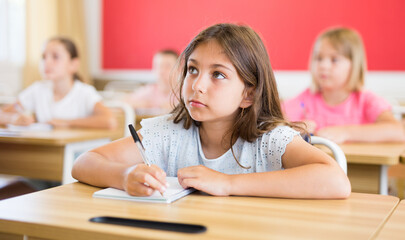 Portrait of focused preteen schoolgirl writing exercises in workbook during lesson in classroom ..