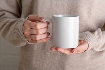 Female hands holding white mug mockup with blank copy space for your advertising text message or promotional content. Girl in beige sweater holding white porcelain coffee mug mock up, close up