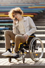 Sad, young disabled guy in a wheelchair talking on the phone. He is sitting in a chair by the stairs.
