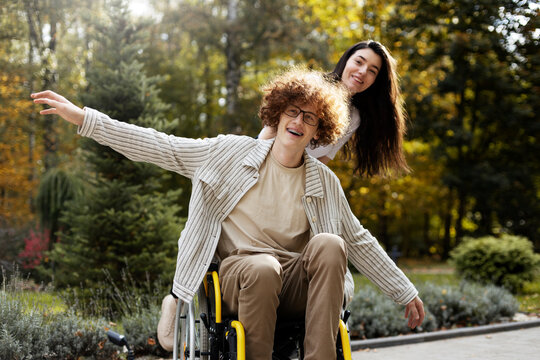 Happy, young man in a wheelchair cheerfully spread his arms to the sides, outdoors. Smiling boy and girl are walking in the park.