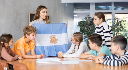 Obraz na płótnie Canvas Group of curious preteen learners and young female teacher with flag of Argentina in classroom of academy. Academic school children learning concept