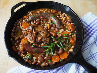French Cassoulet, a meat and beans stew, in a cast iron skillet, on a wooden table with a checkered white and purple dish towel
