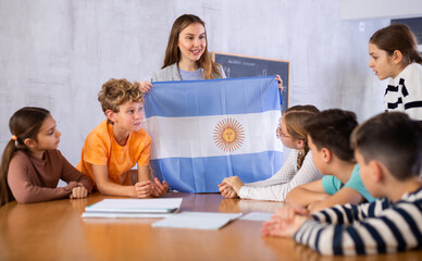 Decent teacher showing Argentina flag to group of preteen schoolchildren in classroom during lesson