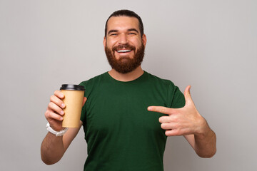 Young smiling bearded man is pointing at the paper coffee cup he is holding over grey background.