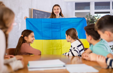 Group of interested preteen learners and young female teacher with flag of Ukraine in classroom of...
