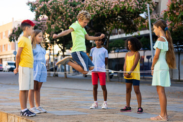 Fototapeta na wymiar Happy preteen kids of different nationalities skipping on chinese jump rope in courtyard on summer day..