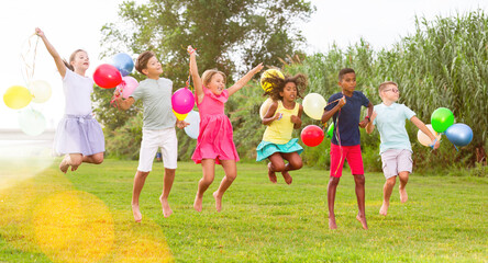 Young joyful boys and girls jumping on lawn with balloons in hands.