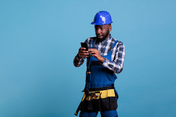Obraz na płótnie Canvas African american builder looking with amazed expression at cell phone screen, wearing coveralls and hard hat. Electrician with tool belt using mobile device against light blue background.