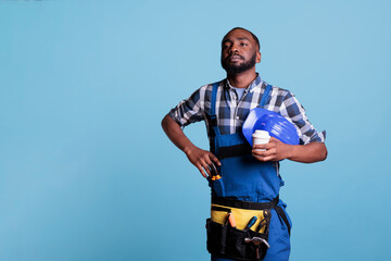 African american builder looking into the distance while resting with a cup of coffee in studio shot against blue background. Construction employee resting before continuing work day.