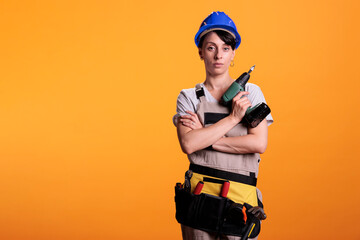 Engieering supervisor holding electric screw gun for construction industry advertisement in studio. Female builder worker in overalls and hard hat standing on yellow background with power drill.