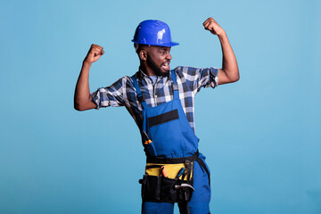 Funny african american construction worker in dungarees showing arm muscles in front of the camera...