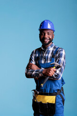 Happy African american builder holding hammer posing in studio against blue background. Workman in hard hat and construction overalls, safety uniform in construction industry.