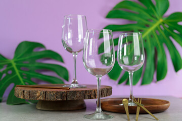 three empty wine glasses on a concrete table with natural wooden planks against a lilac purple background with green monstera leaves decorating the background