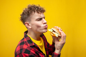 curly guy student eats a burger on a yellow background and shows surprise, happy man with fast food