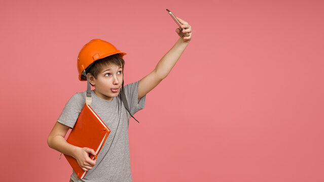 A little builder in a gray T-shirt and a notebook tries to make a good selfie on a pink background