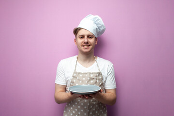 young guy housewife in an apron and a hat holds an empty plate and shows surprise on a pink background