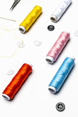 View of black and white buttons, needle with threaded yellow thread, various colors on thread spools on white background