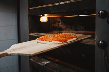 Baker taking hot pizza out of oven with wooden shovel in a bakery