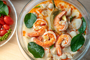 Tom Yam Kung or Tom Yum soup is Thai hot spicy soup shrimp or prawn with lemon grass, lemon, galangal, mushrooms, coconut milk and chili in bowl on wooden table top view, famous Thai food cuisine.