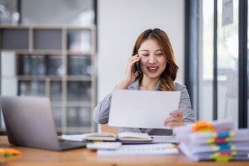Smiling young Asian woman sitting and call phone device, looking at laptop, checking modern apps, texting messages, browsing internet doing shopping relaxing at home.