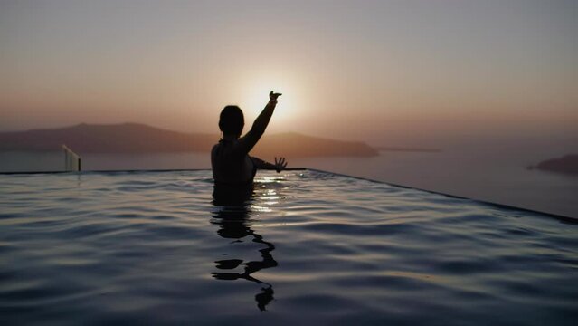 A woman dancing in infinity pool during sunset over sea - Slow motion, Santorini, Greece