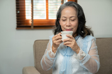 Happy Asian retired woman enjoying her morning tea in the house An elderly woman relaxingly pours and sips hot tea or coffee on the sofa inside her house.