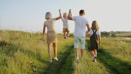 Fototapeta premium Young parents with their son and daughter are walking along the road in the field. Father, son, mother, daughter are walking in the park. Family holding hands. Children with parents walk in nature.