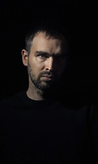 Portrait of a stern man with a small beard looking into the camera. Caucasian man on dark background. Half of his face illuminated. Dramatic portrait of a handsome man.