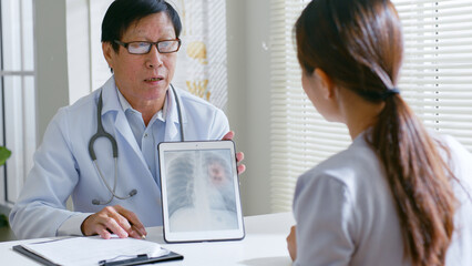 Senior Asian doctor showing lung x-ray test results and describe to a young woman patient in...