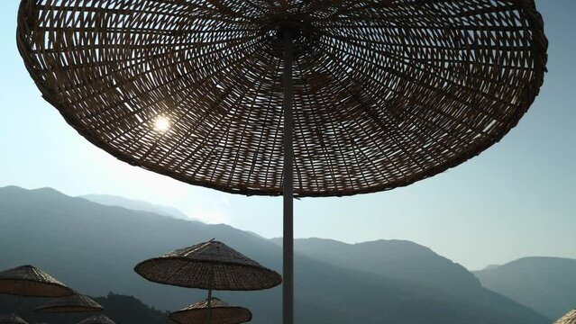 Straw umbrella against blue sky. Close-up. Smooth camera movement from bottom to top. Mountains, beach with holidaymakers in the background. Summer holiday in the Mediterranean