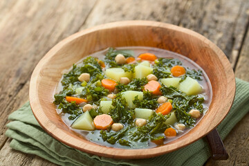 Fresh homemade vegan vegetable soup made of kale, chickpea, carrot, celery, onion and potato served in wooden bowl, photographed on wood (Selective Focus, Focus one third into the soup)