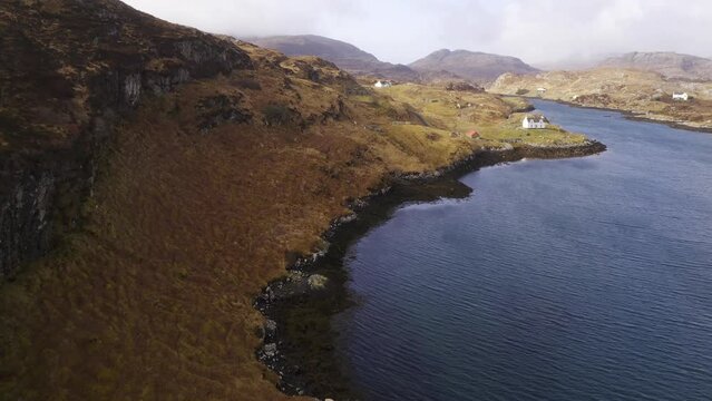 Drone shot of a collection of remote, coastal houses with a Scottish mountain range, moorland and peatland in the background. Filmed on the Outer Hebrides of Scotland.