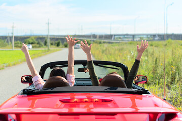 Back view of happy girls driving red cabriolet car with raised hands during vacation road trip having fun together discovering new places. Road trip travel enjoying freedom concept. Selective focus