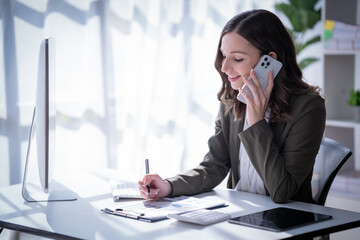 Sharing good business news. Attractive young businesswoman talking on the mobile phone and smiling while sitting at her working place in office and looking at laptop PC.