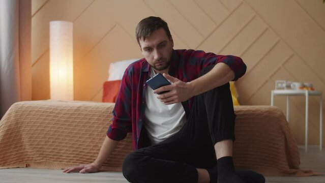A Caucasian angry man in a plaid shirt sits on the bedroom floor by the bed and scrolls the Internet in his smartphone. A floor lamp is turned on in the corner of the room