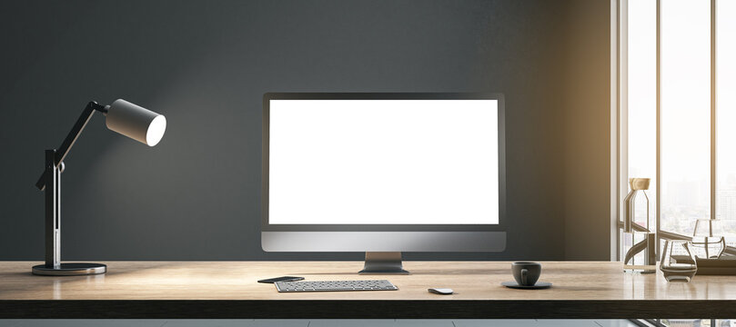 Blank white modern computer monitor with space for your web site or web design on wooden table with coffee cup, keyboard and lamp on dark wall background in sunlit room. 3D rendering, mock up