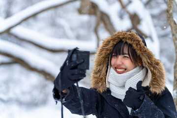 Obraz premium Asian woman using digital camera taking selfie during walking down small town street covered in snow. Attractive girl enjoy travel local village near the mountain in Japan on winter holiday vacation.