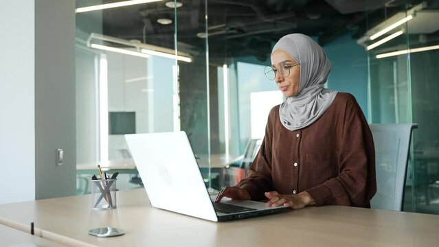 Muslim young businesswoman entrepreneur in hijab feels hot during computer work and turn on remote control air conditioner cooling system or humidifier changing temperature at modern office workplace