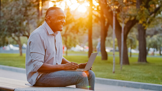Cheerful carefree senior African American man sitting outdoors work with laptop play online game having fun laughing typing on computer. Ethnic mature male middle-aged freelancer working on park bench