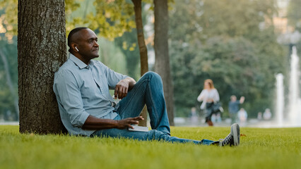 Obraz premium Calm relaxed sad serious middle-aged male listening music with earphones mature adult African American ethnic man in wireless headphones sitting on grass in city park enjoy audio song sound outdoors