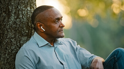 Headshot portrait close up elderly middle-aged adult African American man businessman relaxing sitting under tree in city park work break listening music audio song sound with wireless headphones