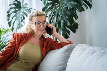Happy senior woman talking through smart phone leaning on sofa at home