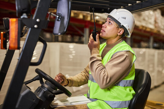 Side view portrait of female worker wearing hardhat while operating forklift truck in warehouse and talking to portable radio