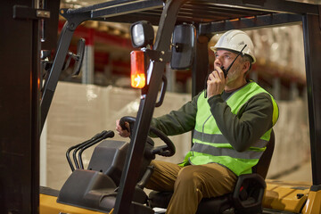 Portrait of male worker driving forklift truck in warehouse and talking to portable radio