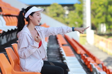 Portrait of happy and excited young female asian sport fan cheering and watching the match with her finger pointing, she sitting amongst the rows of empty seats on stadium while celebrate winner team