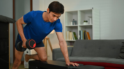 Athlete man doing exercise with dumbbell leaning on sports bench in living room. Fitness and bodybuilding concept