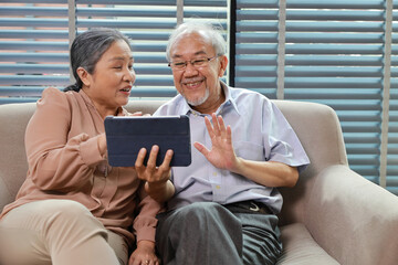 Obraz premium Happy smiling asian senior couple sitting on sofa and using tablet while online video call with friend or relative cousin at home living room. Internet information technology and lifestyle concept.