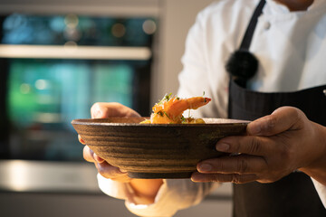 Chef's hand holds the cooked shrimp in bowl with blur oven background behind.